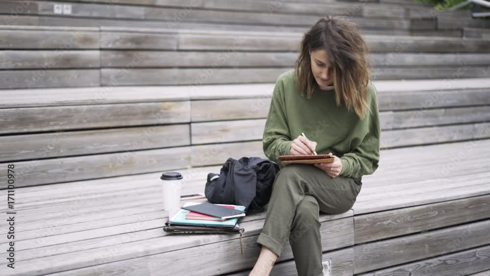 Pretty young woman is sitting on wooden stairs outdoors and drawing at her tablet computer with a special pen. Locked down real time establishing shot Pretty young woman is sitting on wooden stairs outdoors and drawing at her tablet computer with a special pen. Locked down real time establishing shot