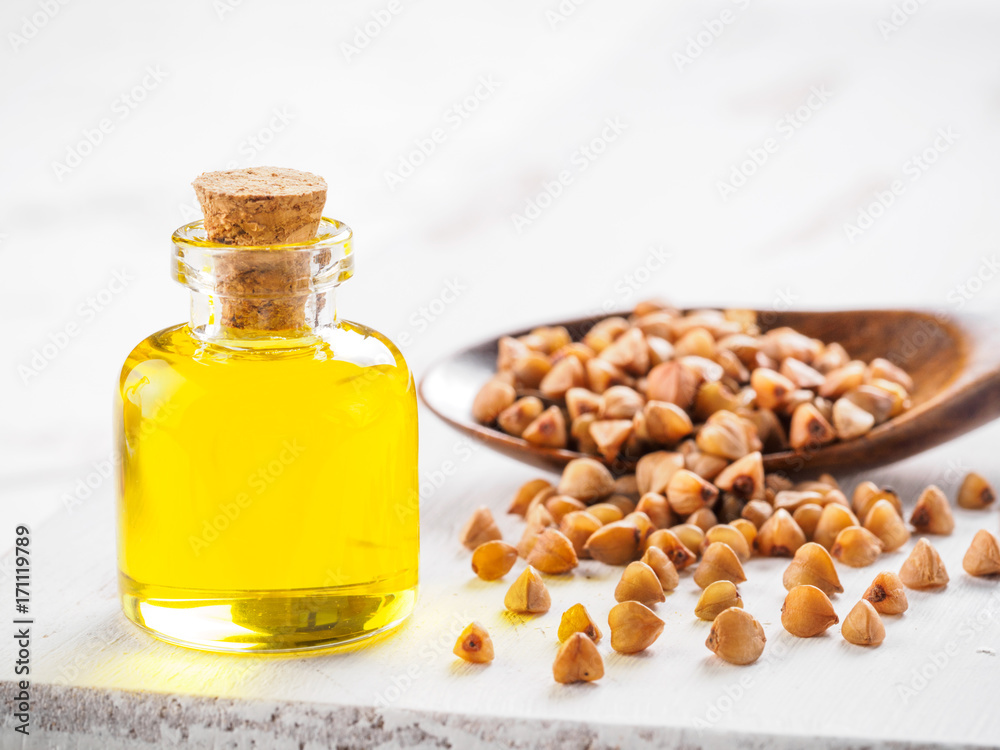 Brown buckwheat in spoon and buckwheat oil in glass bottle on white wooden background. Copy space. Brown buckwheat in spoon and buckwheat oil in glass bottle on white wooden background. Copy space.