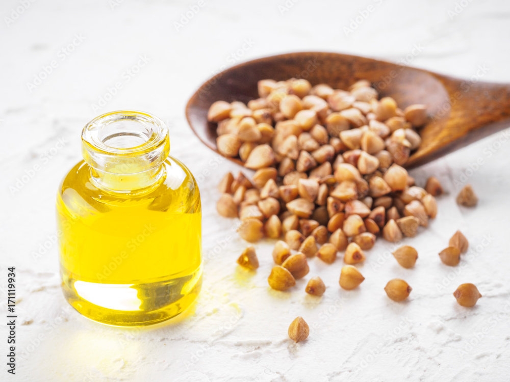 Brown buckwheat in spoon and buckwheat oil in glass bottle on white wooden background. Copy space.