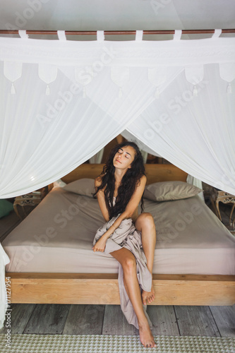 Morning of beautiful young woman pleasuring in bed. Room in Bali style with wooden furniture and white canopy © Oleg Breslavtsev