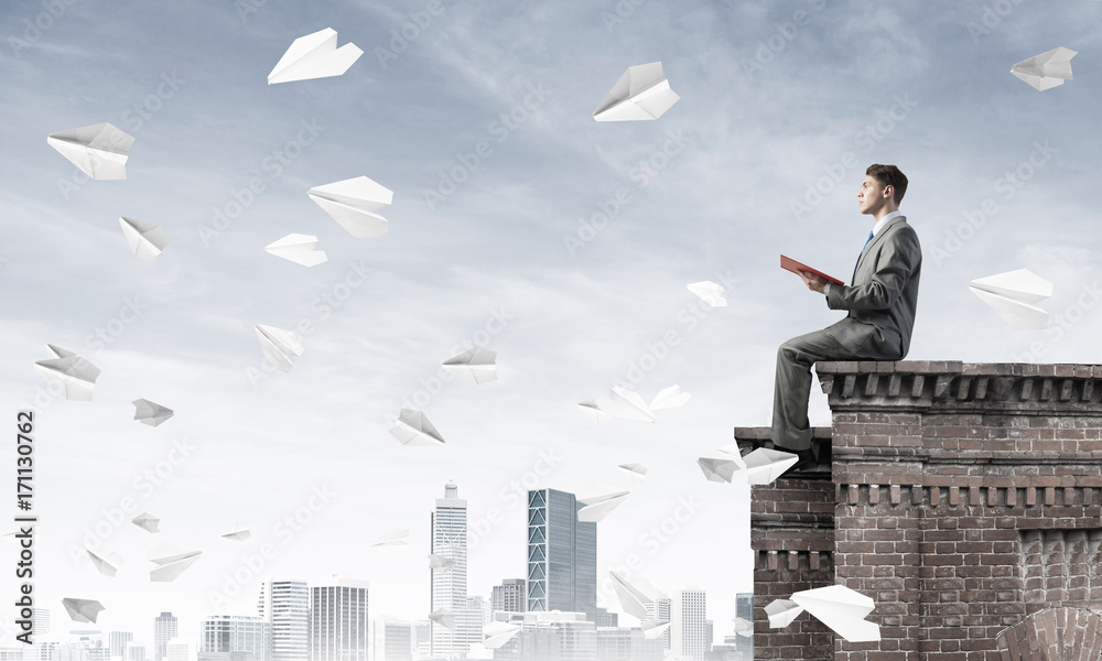 Man on roof edge reading book and cityscape at background