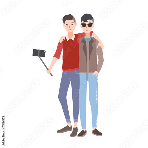 Two young men dressed in stylish clothing standing together, smiling and making selfie photo using monopod with smartphone