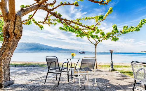 Street outdoor cafe on Embankment of Corinth Gulf beneath the tree. View at solitary vessel on horizon. Beautiful sea view on breathtaking vista, amazing spring scenery. Fresh blue sky over scene. photo
