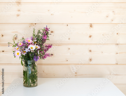 Bouquet of purple and white spring flowers, including daisies, in glass jar on white table against pine board wall