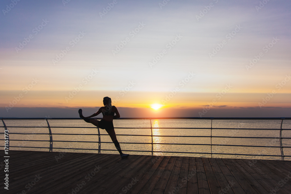 Silhouette of young beautiful athletic girl with long blond hair in headphones listening to music and doing stretching at sunrise over the sea, back view