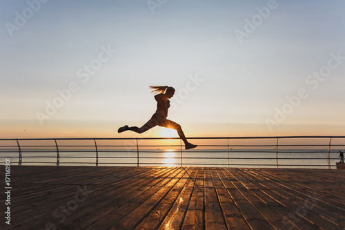 Silhouette of a young beautiful athletic girl with long blond hair in headphones, who listens to music and runs at dawn over the sea