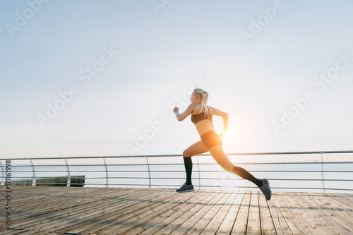 Young beautiful athletic girl with long blond hair in headphones listening to music and running at sunrise over the sea