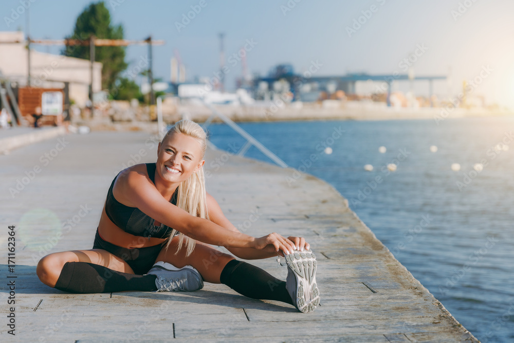 Young beautiful athletic girl with long blond hair in headphones and mobile phone on her hand listening to music and doing stretching at sunrise over the sea, close-up