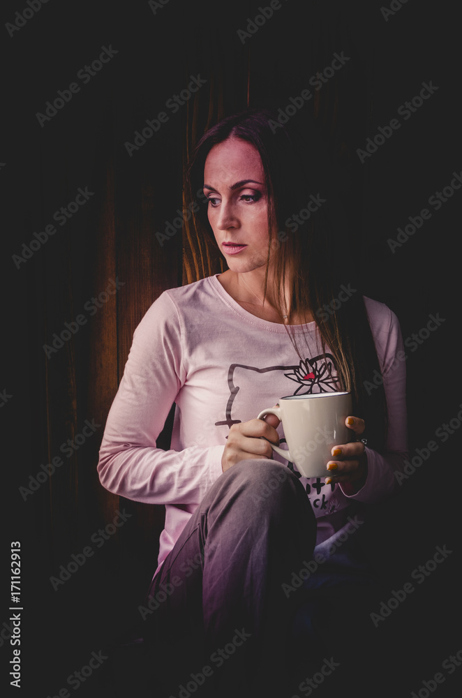 Beautiful young woman drinking coffee and looking through window while sitting at windowsill at home