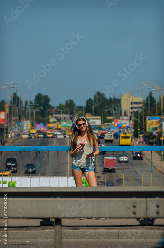Beautiful stylish girl student stands on the car bridge with a beautiful view of the city and drinks music
