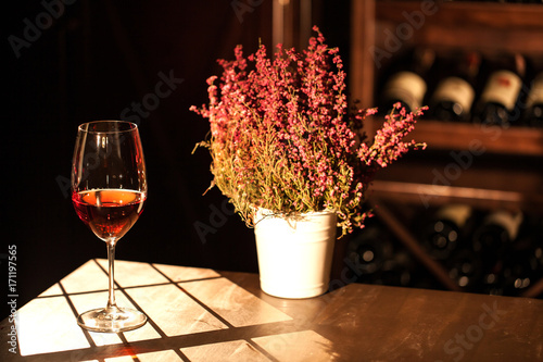 Fototapeta Horizontal romantic composition of the glass of wine and flower pot placed on the table in the winehouse