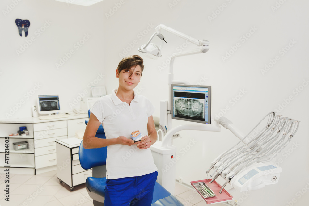 Obraz premium Woman dentist with short hair in a white shirt and blue trousers holds dentures in the dental office