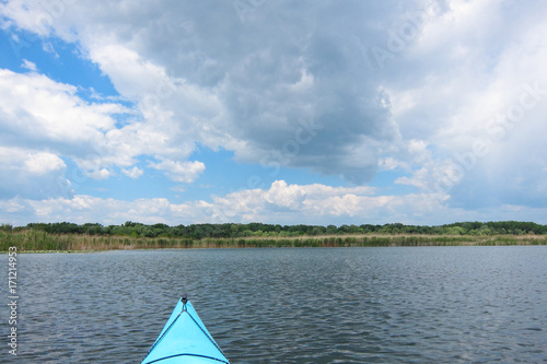 Fototapeta Naklejka Na Ścianę i Meble -  Bow of blue kayak. View over lake with green trees and cloudy sky from blue kayak. Kayaking in calm spring day. Reserve on Danube river delta