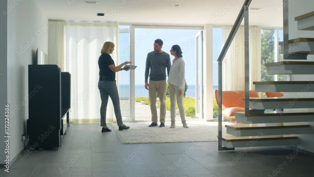 Professional Real Estate Agent Shows Stylish Modern House to a Beautiful Young Couple Who are in the Market for Purchasing/ Renting New Home. House Has Floor to Ceiling Windows and Seaside View. Professional Real Estate Agent Shows Stylish Modern House to a Beautiful Young Couple Who are in the Market for Purchasing/ Renting New Home. House Has Floor to Ceiling Windows and Seaside View.