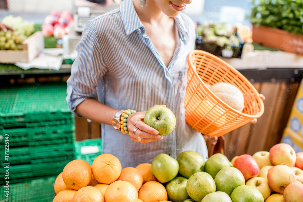 Woman choosing a fresh apples standing with basket at the food market with fruits and vegetables