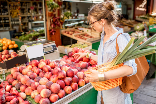 Photos Young woman choosing a fresh peach standing with basket at the food market in Fr