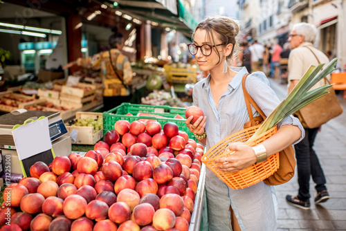 Canvas Print Young woman choosing a fresh peach standing with basket at the food market in Fr