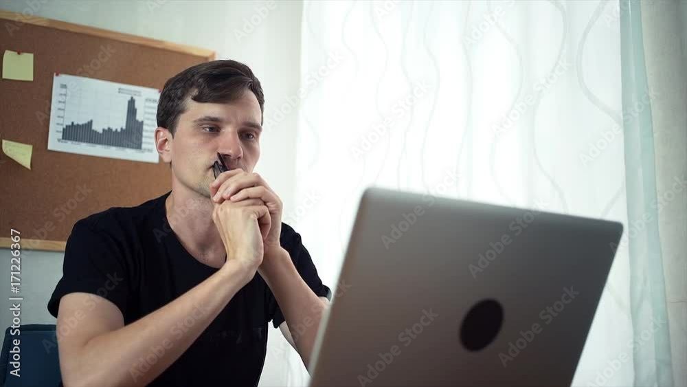 Freelancer holds in hands glasses, look at the laptop screen. A board with stickers and graphs on the wall behind Freelancer holds in hands glasses, look at the laptop screen. A board with stickers and graphs on the wall behind