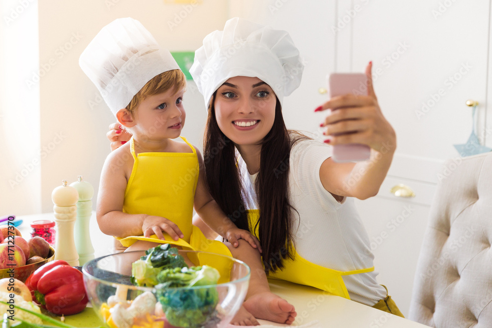 The young smiling cook mother sitting with her little son in the kitchen near vegetables and doing selfie-photo on the phone The young smiling cook mother sitting with her little son in the kitchen near vegetables and doing selfie-photo on the phone
