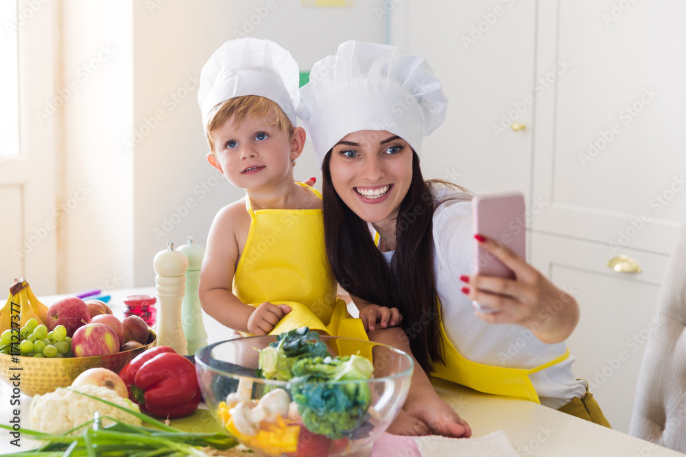 The young smiling cook mother sitting with her cute little son in the kitchen near vegetables and doing selfie-photo on the phone The young smiling cook mother sitting with her cute little son in the kitchen near vegetables and doing selfie-photo on the phone