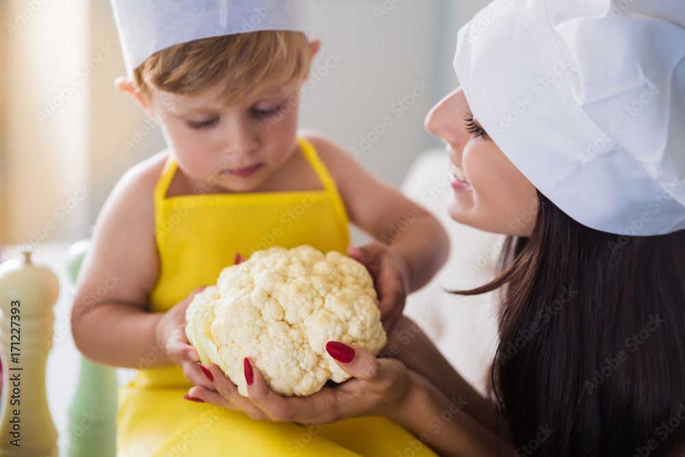 Portrait of young smiling cook mother with her cute little son in the kitchen, they keeping cauliflower in hands Portrait of young smiling cook mother with her cute little son in the kitchen, they keeping cauliflower in hands