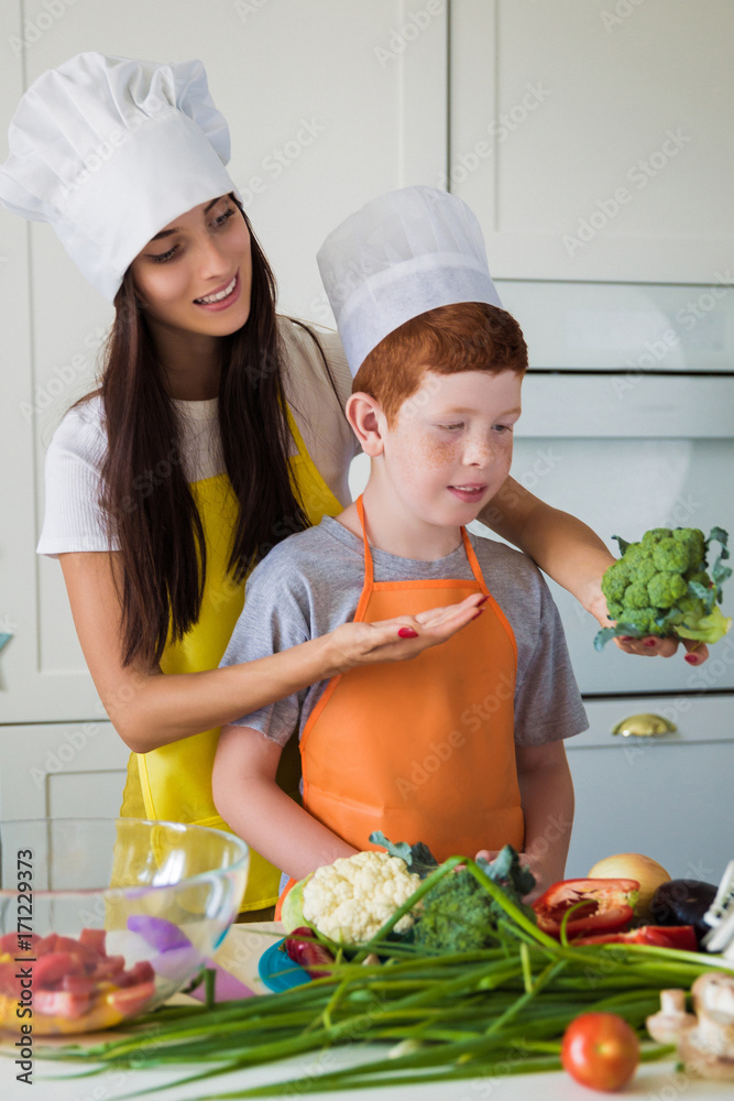 The young loving mother with her son standing in cook form in the kitchen and showing broccoli to her son