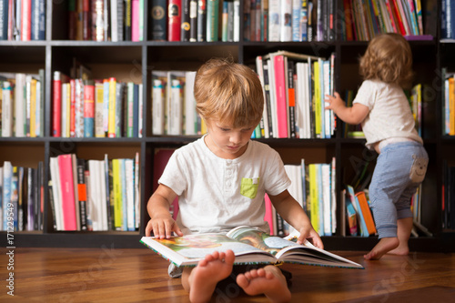Cute boy and his little sister  reading a book together  near bookshelf at home 