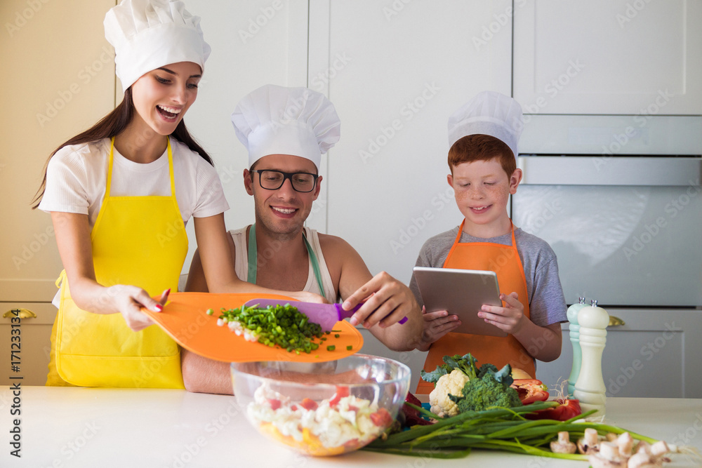 The young family in cook form in the kitchen, mother and father cooking salad, son watching tablet The young family in cook form in the kitchen, mother and father cooking salad, son watching tablet