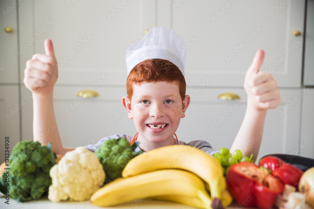 Portrait of young joyful cook boy who sitting by the table with fruits and vegetables in the kitchen and showing thumbs up Portrait of young joyful cook boy who sitting by the table with fruits and vegetables in the kitchen and showing thumbs up