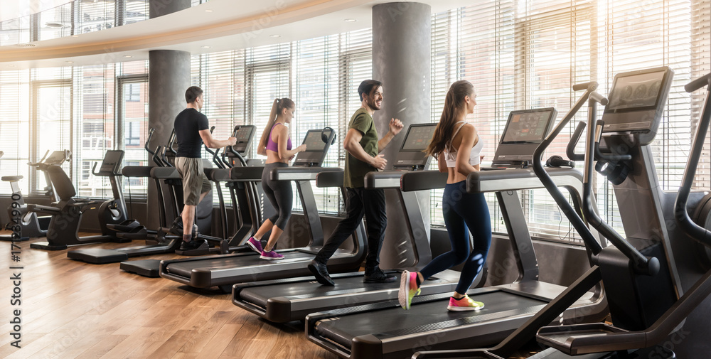 Fototapeta premium Grupa czterech osób, mężczyzn i kobiet, biegających na bieżniach w nowoczesnej i oświetlonej sali fitness