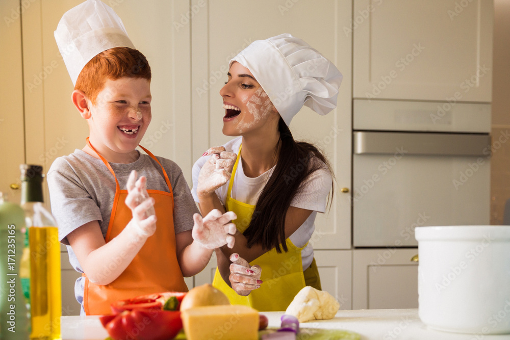 The young cook woman and her son in cook form kneading the dough and they dirtying of flour each other The young cook woman and her son in cook form kneading the dough and they dirtying of flour each other