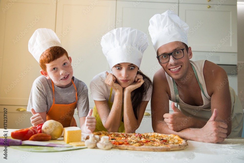 The young happy family in cook form standing in the kitchen and looking at pizza and father and son showing thumbs up The young happy family in cook form standing in the kitchen and looking at pizza and father and son showing thumbs up