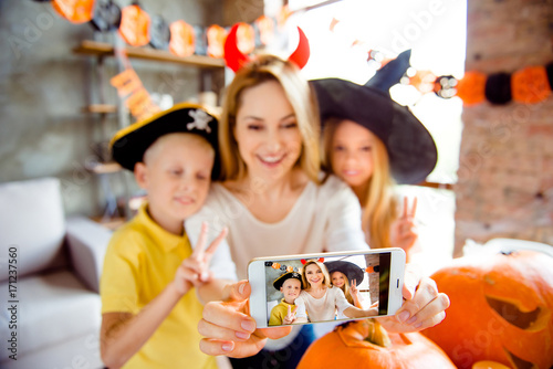 Kids say cheers! Dressed in spooky halloween costumes headwear mum and her cheerful kids are bonding embracing, lady is taking selfie, making memories, siblings gesturing, creepy decorations
