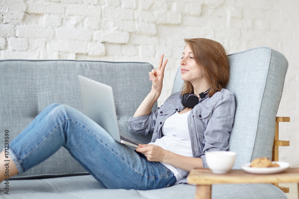 Candid shot of cheerful young European woman in jeans sitting on  comfortable sofa with notebook pc