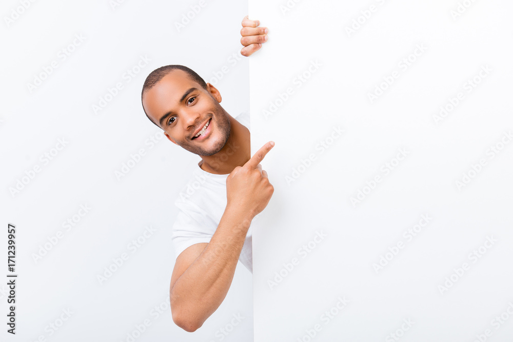 Happy amazed young guy is popping out from white blank banner, pointing at it with finger, wearing white t shirt, on white background Happy amazed young guy is popping out from white blank banner, pointing at it with finger, wearing white t shirt, on white background