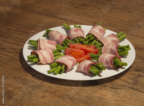 green bean rolls in bacon on white plate with sliced tomato on wooden table background