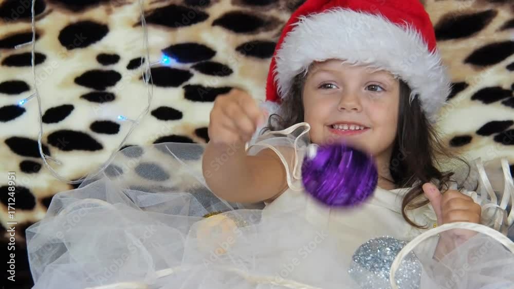 A beautiful little girl is twirling a New Year ball. The child in the cap of santakluus plays with New Year's toys. A beautiful little girl is twirling a New Year ball. The child in the cap of santakluus plays with New Year's toys.