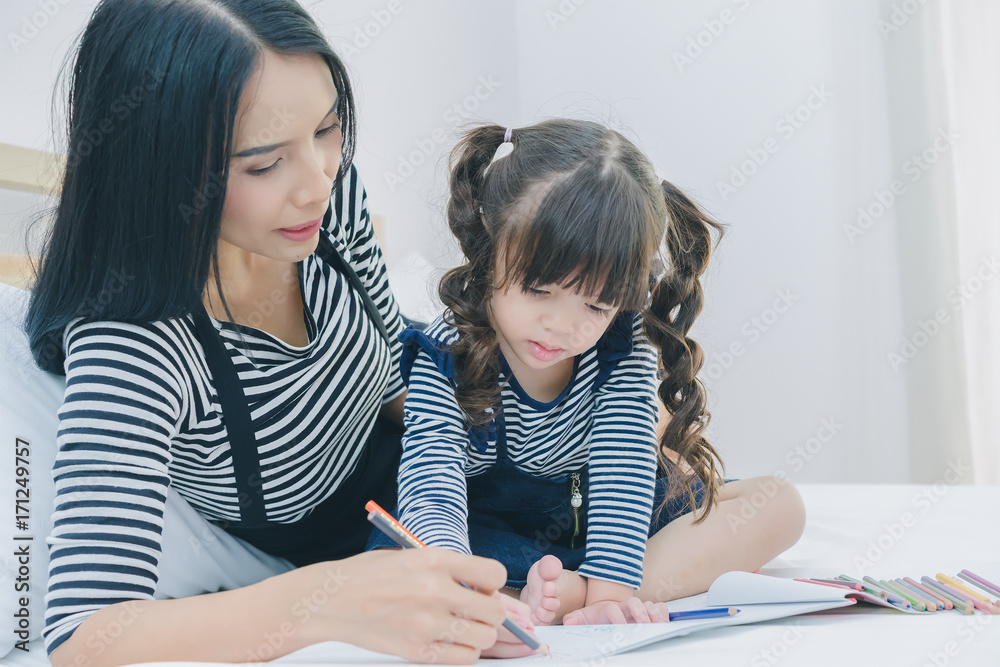 A little cute girl enjoy drawing into the book by colorful pastel with mother on the white bed in the bedroom, education time at home A little cute girl enjoy drawing into the book by colorful pastel with mother on the white bed in the bedroom, education time at home