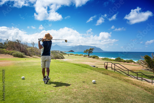 Golfer with straw hat teeing off with driver from ocean side par 4 hole