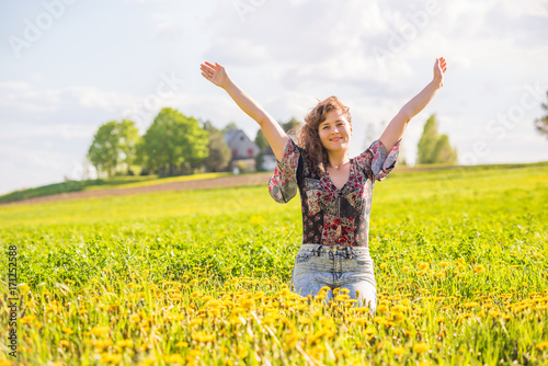Young happy smiling woman sitting in yellow dandelion flower farm field during sunset by countryside rural house in Ile D'Orleans, Quebec, Canada raising hands