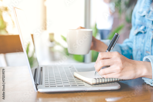A woman's hand working on a notebook computer and writing on a notepad with a pen in the office.