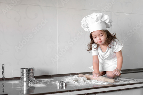Cute little girl in apron and chef hat is kneading the dough and smiling while baking