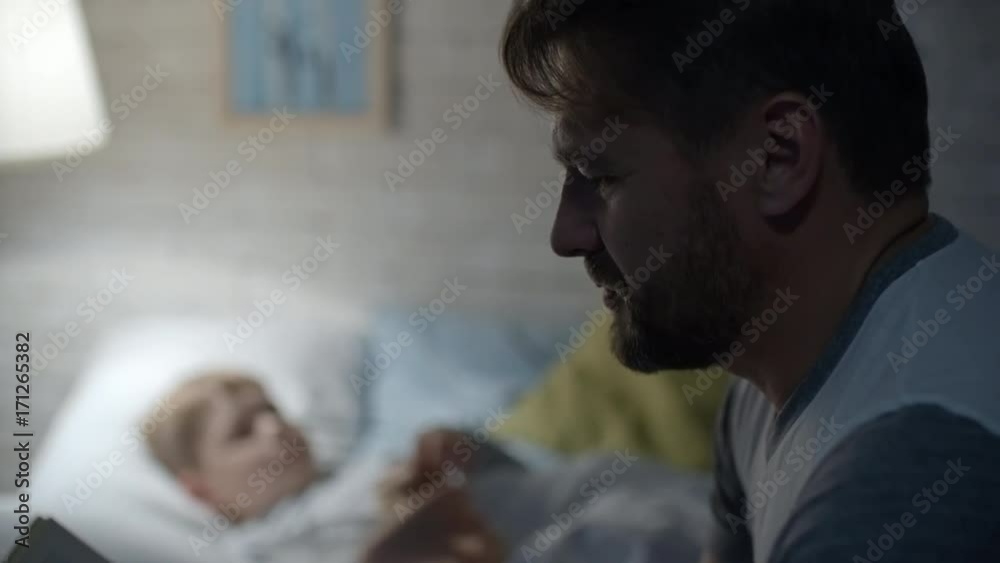 Tilt up of loving father reading fairytale to little son lying in bed and playing with toy before bedtime in cozy bedroom Tilt up of loving father reading fairytale to little son lying in bed and playing with toy before bedtime in cozy bedroom