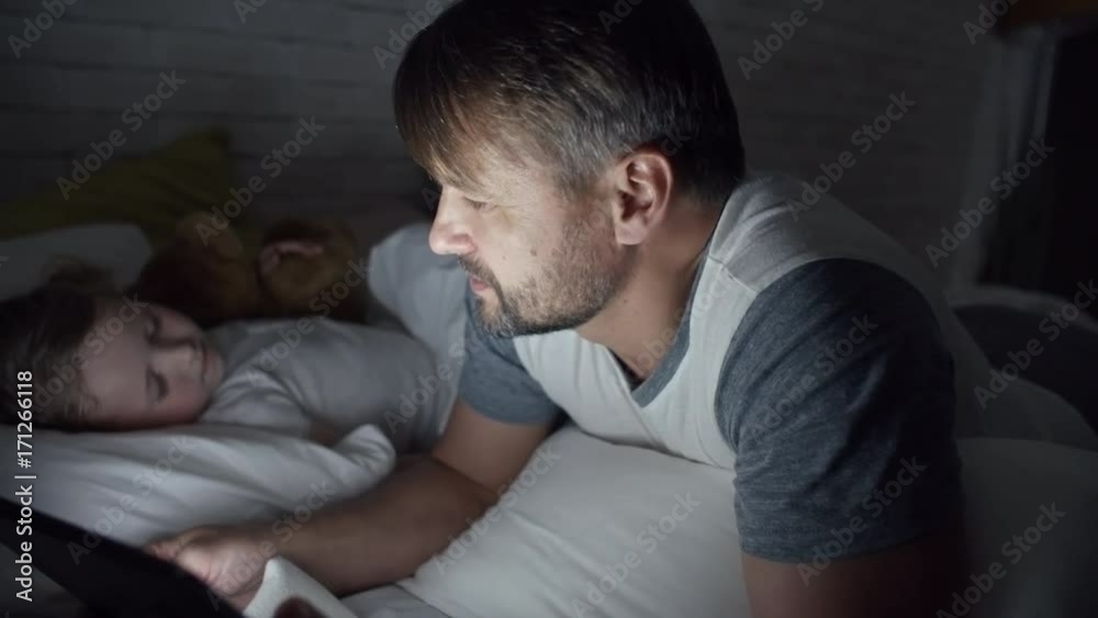 PAN of bearded father lying on bed and reading story on tablet to adorable girl sleeping beside PAN of bearded father lying on bed and reading story on tablet to adorable girl sleeping beside