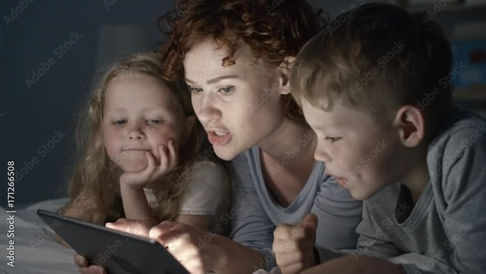 PAN of beautiful mother with short curly hair lying in bed, chatting with adorable boy and girl and looking at tablet PAN of beautiful mother with short curly hair lying in bed, chatting with adorable boy and girl and looking at tablet