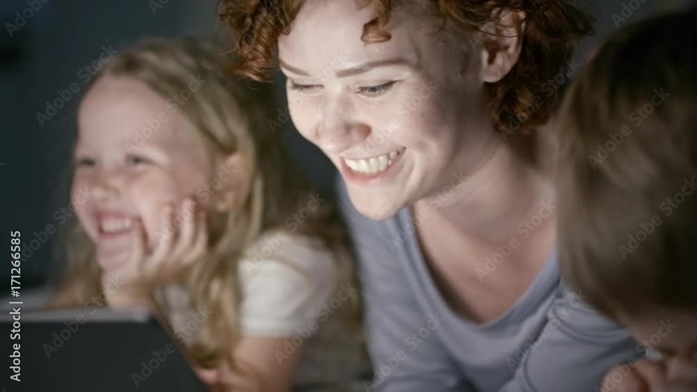 Tilt down with slow motion of beautiful young mother with short curly hair lying in bed with adorable children and reading aloud digital book on tablet, then joking and laughing together