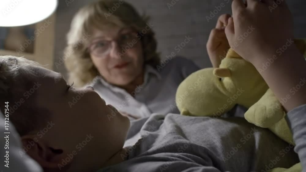 PAN of happy little boy with plush toy lying in bed and chatting with cheerful grandmother in glasses PAN of happy little boy with plush toy lying in bed and chatting with cheerful grandmother in glasses
