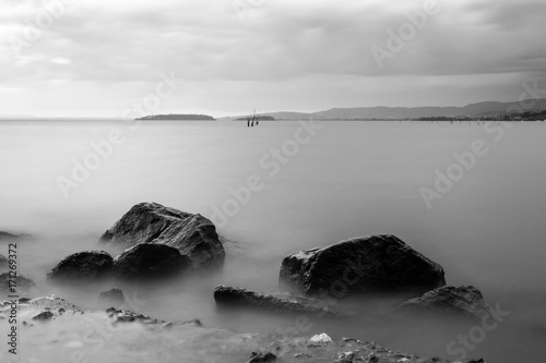A minimalist, long exposure photo of some big rocks in a lake, with perfectly still water and cloudy sky