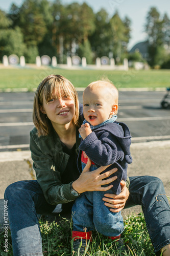 Happy young mother with her son sit on the grass, smile and hug.Outdoors healthy child activity, active lifestyle and having fun on family summer vacation with son at the playground.