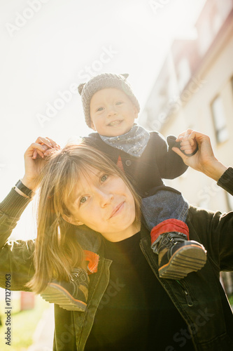 Happy young mother with her son on her shoulder, enjoying the sunshine, they smile and laugh.Outdoors healthy child activity, active lifestyle and having fun on family summer vacation with son at the 